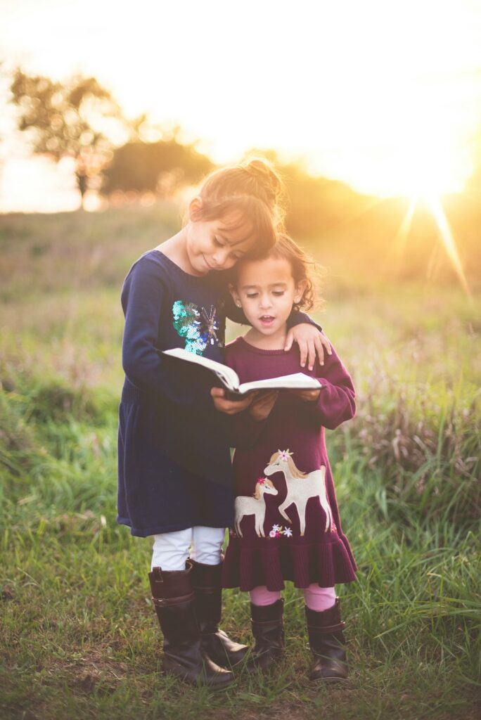 Two young girls standing in a grassy field reading a book together, with sunlight in the background.